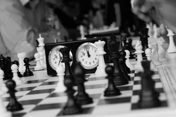 Black and white image of chess board in a middle game with analog chess clock in the background.