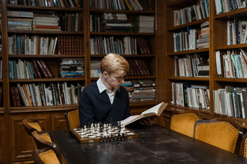 A young man sits in a library reading a chess book and recreating the chess position on a board.