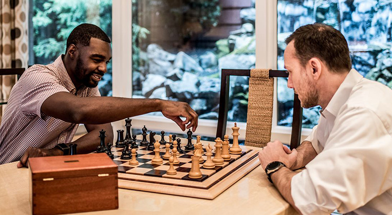 two men playing chess on a table with the leftmost man reaching for a chess piece