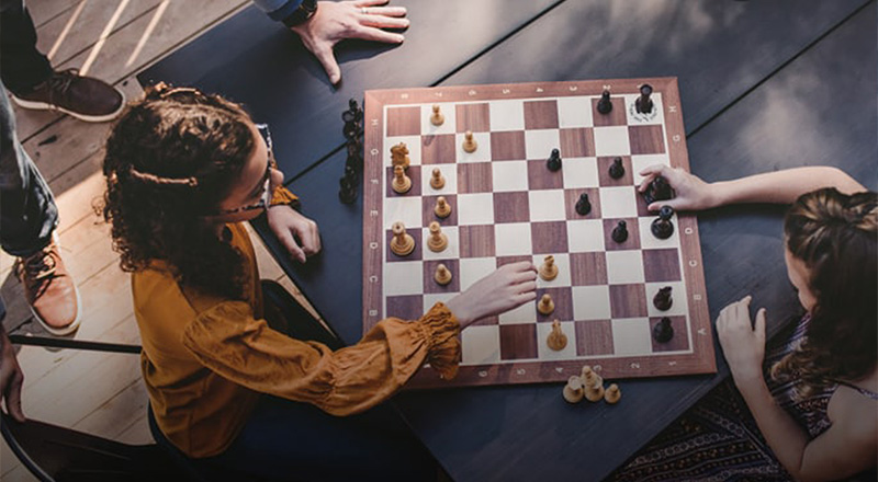 two women playing chess on a large wooden chessboard in a park